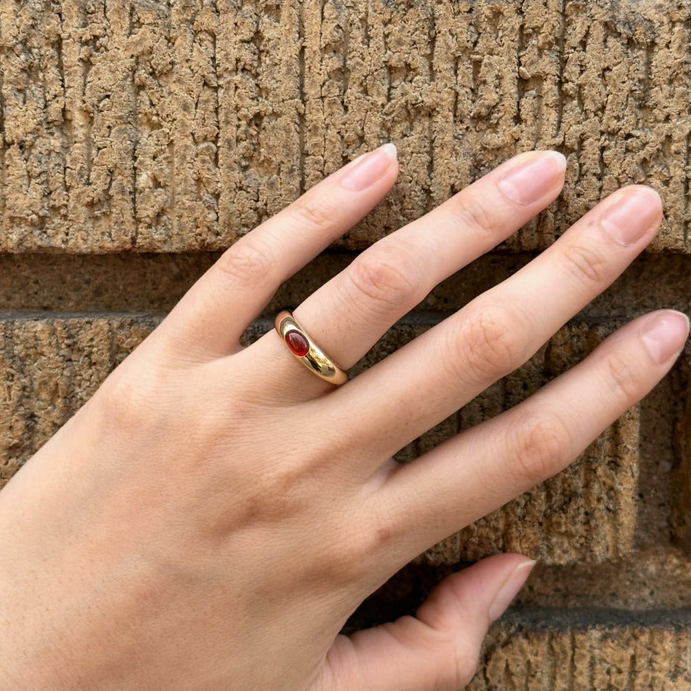 Hand wearing a gold ring with a red stone against a textured stone wall.