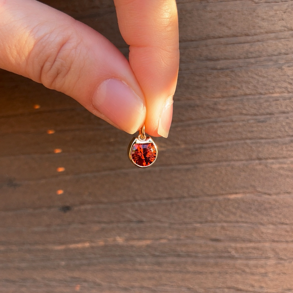 A close-up of a hand holding a small gold cat face silhouette pendant with a red stone against a brown wooden background.