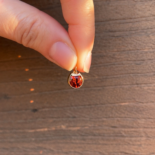 A close-up of a hand holding a small gold cat face silhouette pendant with a red stone against a brown wooden background.
