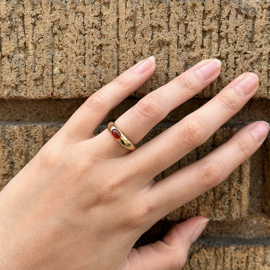 Hand wearing a gold ring with a red stone against a textured stone wall.